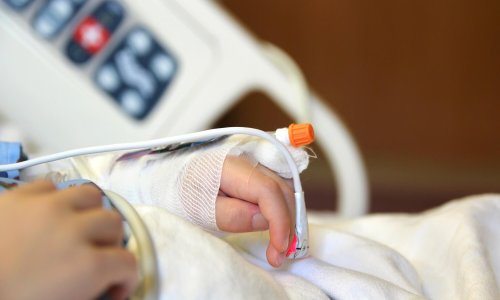 A child is laying on a hospital bed with his arm and hand raised above his head with an I.V. and bandage on it.